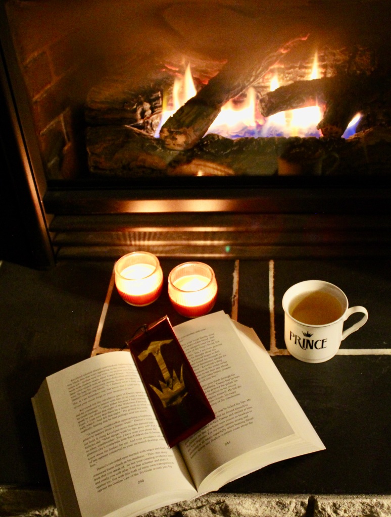 An open book is placed before a fireplace with a bright fire. There is a bookmark with a hammer and crown symbol in the book. There is a teacup with the word prince to the right of the book. Two orange jar candles are burning above the book.